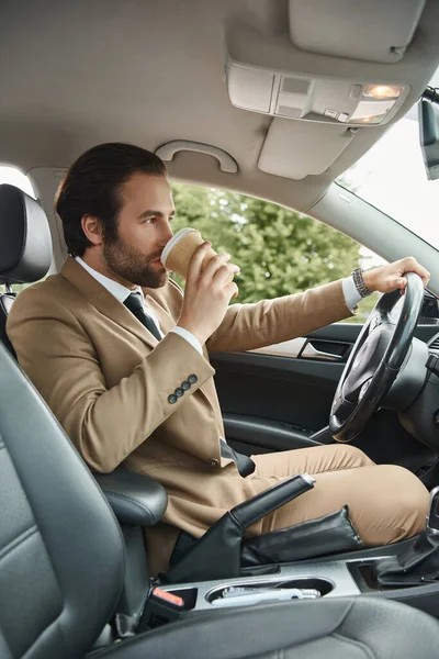 Stylish bearded businessman in beige suit drinking coffee to go and driving car on urban street — Stock Photo