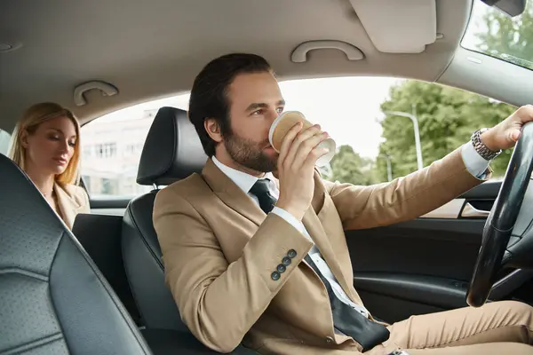 Hombre con estilo en ropa formal beber café y conducir un coche con mujer de negocios rubia en el asiento trasero - foto de stock