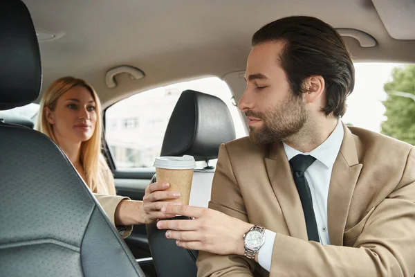 Blonde et élégante femme d'affaires donnant du café pour aller à l'homme élégant sur le siège des conducteurs dans une voiture de luxe — Photo de stock