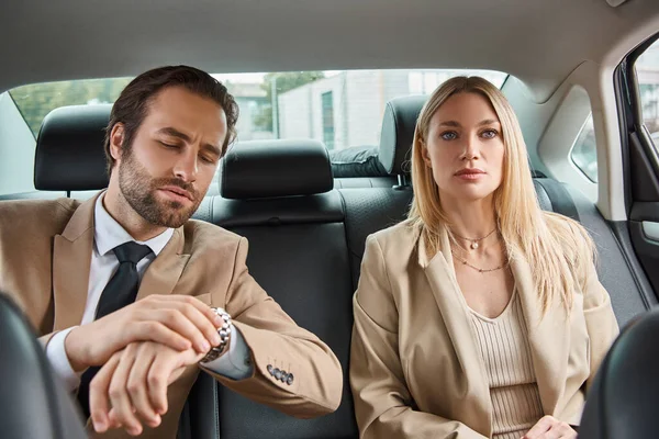 Homme d'affaires inquiet en tenue formelle regardant montre-bracelet pendant le voyage en voiture près de femme blonde — Photo de stock