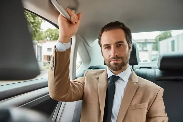 Уверенный и красивый бизнесмен в бежевом элегантном костюме смотрит на камеру в роскошном автомобиле на улице — стоковое фото