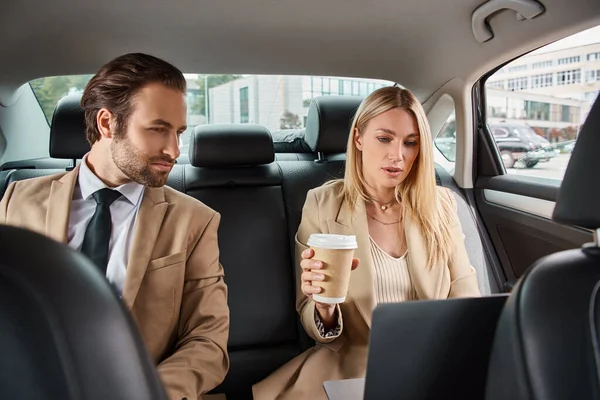 Donna bionda con caffè per andare e laptop vicino a bell'uomo d'affari in abbigliamento formale che viaggia in auto — Foto stock