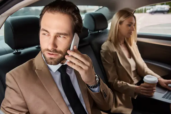 Businessman in formal wear talking on smartphone near woman with coffee to go and laptop in car — Stock Photo