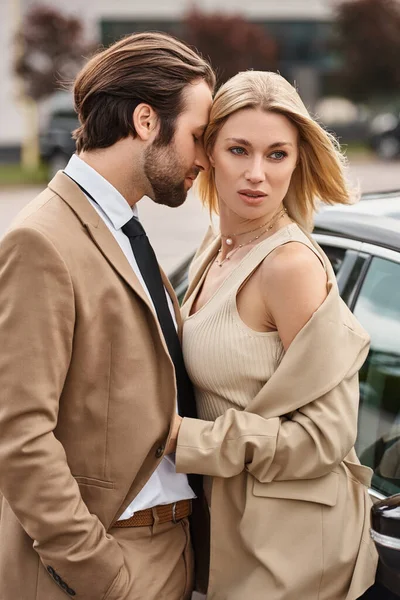 Encantadora mujer de negocios rubia abrazando hombre elegante cerca de coche en la calle, atracción y amor - foto de stock
