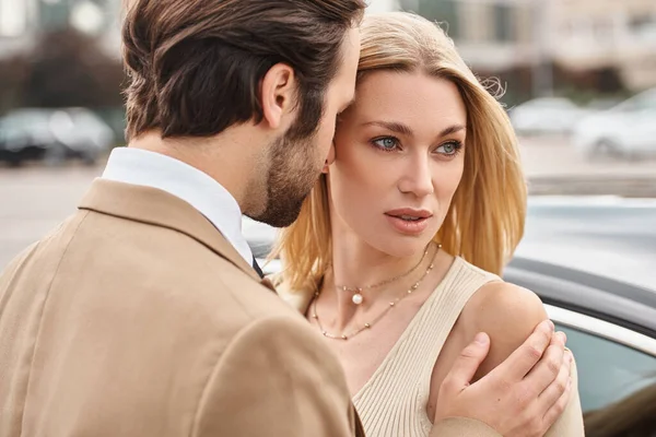 Businessman embracing sensual blonde woman in formal wear near car on urban street, attraction — Stock Photo