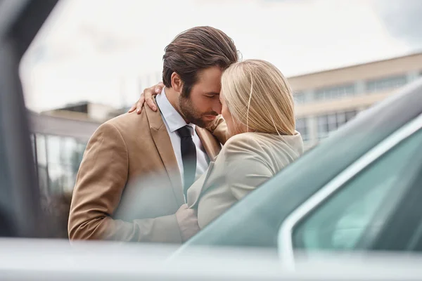 Elegant business couple in formal wear embracing while standing near car on urban street, romance — Stock Photo