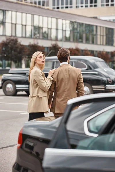 Attractive blonde businesswoman in formal wear touching shoulder of elegant man near car on street — Stock Photo