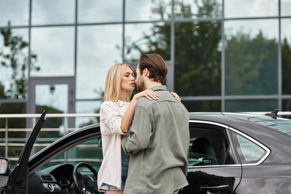 Seductive blonde woman and trendy bearded man in stylish casual clothes embracing near car — Stock Photo