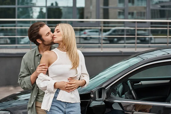 Бородатый мужчина обнимает и целует соблазнительную стильную блондинку возле современного автомобиля на городской улице — стоковое фото