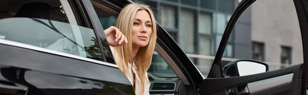 Modern blonde woman in casual clothes sitting in car and looking away on urban street, banner — Stock Photo