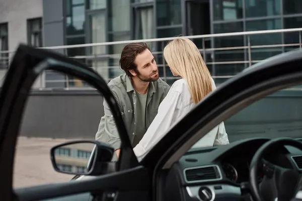 Красивый мужчина смотрит на соблазнительную блондинку возле современного автомобиля на городской улице, влюбленность — стоковое фото
