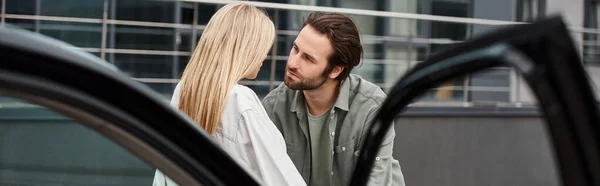 Trendy man looking at stylish blonde woman near modern car on urban street, horizontal banner — Stock Photo