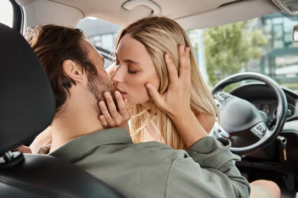 Charming blonde woman kissing man on drivers seat in modern car on city street, urban romance — Stock Photo