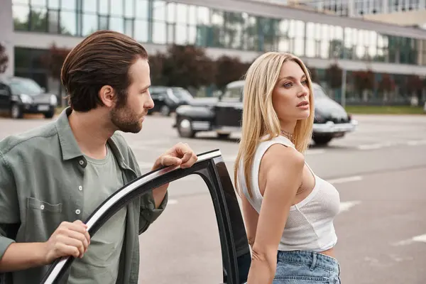 Handsome bearded man looking at alluring blonde woman near car on modern urban street, affection — Stock Photo