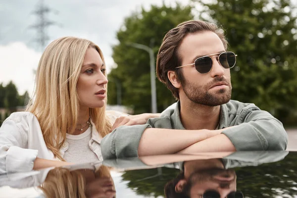 Homme à la mode dans les lunettes de soleil regardant loin près sensuelle femme blonde près de la voiture dans la rue, histoire d'amour — Photo de stock