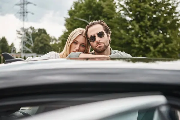 Модный мужчина в солнечных очках и чувственная блондинка, смотрящая на камеру возле машины на улице, история любви — стоковое фото