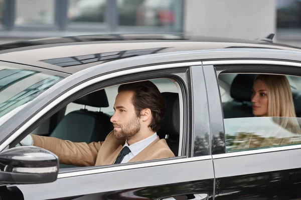 Elegante uomo barbuto in abbigliamento formale guida auto con attraente donna d'affari bionda sul sedile posteriore — Foto stock