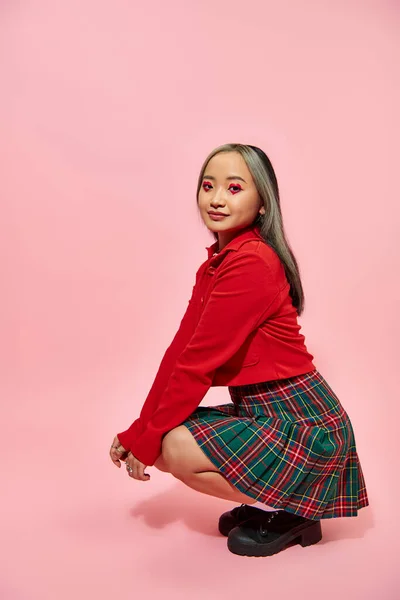Longitud completa de la joven mujer asiática con el corazón en forma de maquillaje de ojos y falda a cuadros sentado en rosa - foto de stock