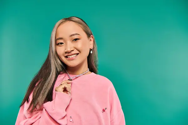 Portrait of cheerful asian woman in pink sweatshirt pulling heart shaped necklace on blue backdrop — Stock Photo