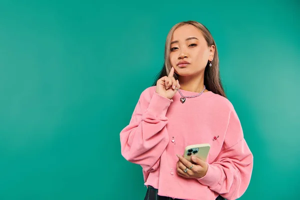 Portrait of pensive young asian woman in pink sweatshirt using smartphone on turquoise backdrop — Stock Photo