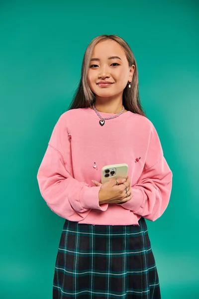 Портрет счастливой молодой азиатки в розовой толстовке и клетчатой юбке с помощью смартфона на бирюзе — стоковое фото