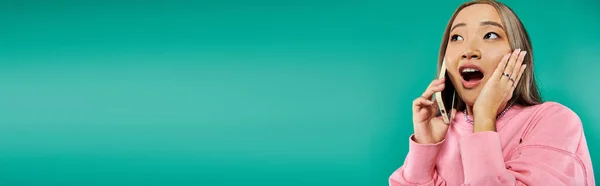 Portrait of shocked young asian woman in pink sweatshirt talking on smartphone on turquoise, banner — Stock Photo