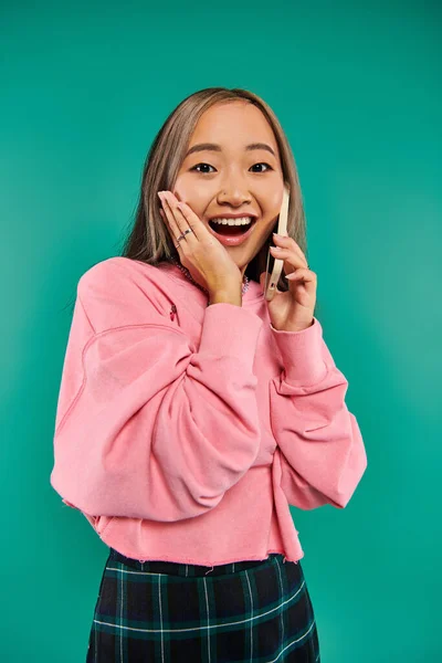 Retrato de mujer asiática joven asombrada en sudadera rosa hablando en smartphone sobre fondo turquesa - foto de stock