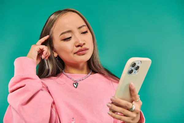 Portrait of thoughtful young asian woman in pink sweatshirt and plaid skirt using smartphone on blue — Stock Photo