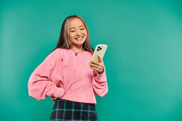 Portrait of positive young asian woman in pink sweatshirt and plaid skirt using smartphone on blue — Stock Photo