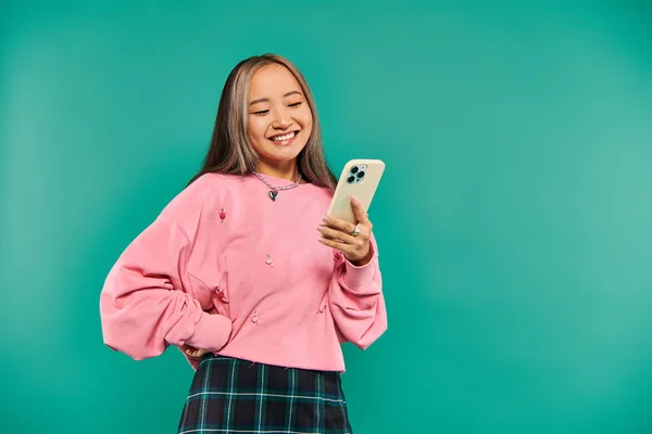 Porträt einer positiven jungen Asiatin in rosa Sweatshirt und kariertem Rock mit Smartphone auf blau — Stockfoto