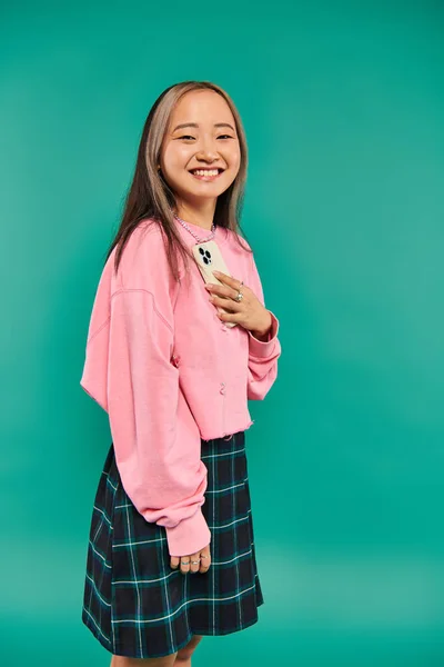 Portrait of positive young asian woman in pink sweatshirt and plaid skirt holding smartphone on blue — Stock Photo