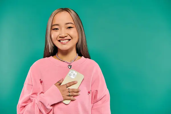 Joyful young asian woman in sweatshirt and plaid skirt holding smartphone on turquoise backdrop — Stock Photo