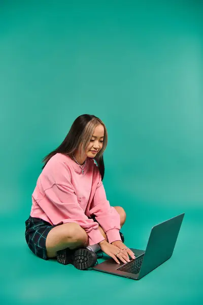 Young asian woman in sweatshirt and plaid skirt sitting and using laptop on turquoise backdrop — Stock Photo