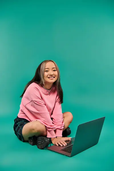 Happy asian woman in sweatshirt and plaid skirt sitting and using laptop on turquoise backdrop — Stock Photo
