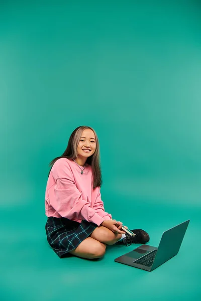 Happy asian woman in sweatshirt and plaid skirt holding phone and using laptop on turquoise backdrop — Stock Photo