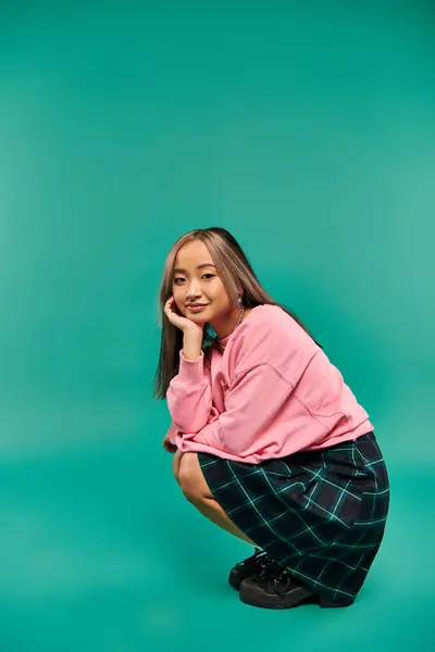 In voller Länge erfreut junge asiatische Frau in rosa Sweatshirt und kariertem Rock sitzt auf rosa Hintergrund — Stockfoto