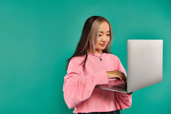 Happy asian woman in pink sweatshirt using laptop while standing on turquoise backdrop, networking — Stock Photo