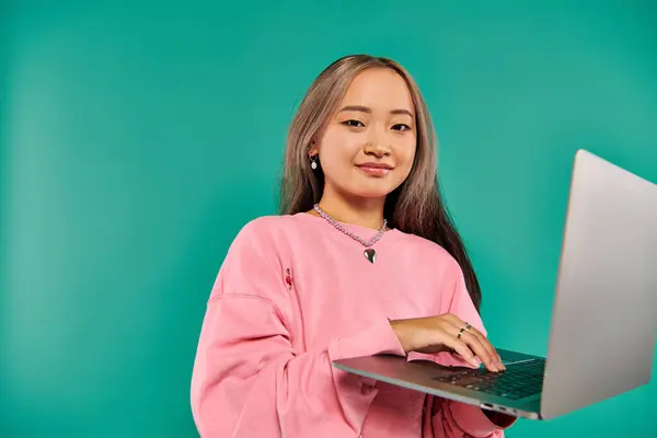 Pleased asian woman in pink sweatshirt using laptop while standing on turquoise backdrop, networking — Stock Photo