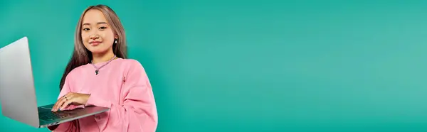 Mujer asiática feliz en sudadera rosa usando el ordenador portátil mientras está de pie sobre el telón de fondo turquesa, bandera - foto de stock