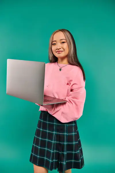 Feliz asiático chica en rosa sudadera usando portátil mientras de pie en turquesa telón de fondo, la creación de redes - foto de stock