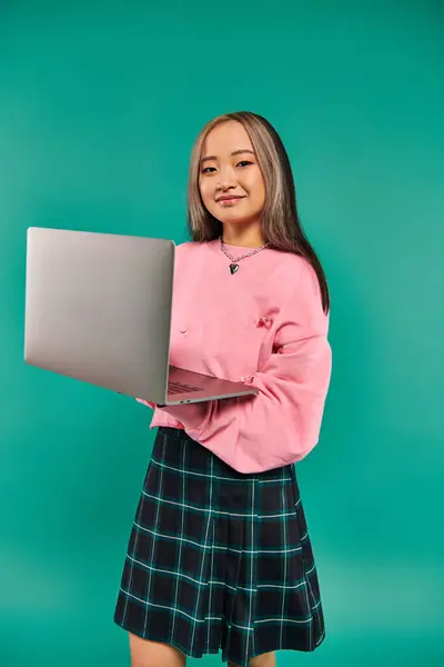 Happy asian girl in pink sweatshirt using laptop while standing on turquoise backdrop, networking — Stock Photo