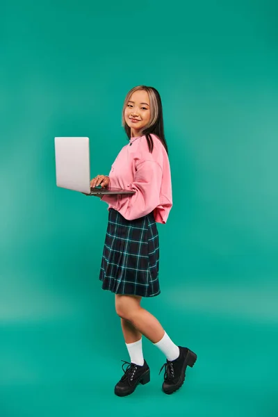 Pleased asian girl in pink sweatshirt using laptop while standing on turquoise backdrop, networking — Stock Photo