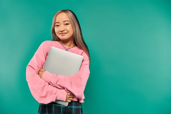 Pleased asian girl in pink sweatshirt holding laptop while standing on turquoise background — Stock Photo