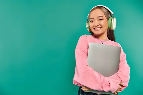 Joyful asian girl in wireless headphones holding laptop while standing on turquoise backdrop — Stock Photo
