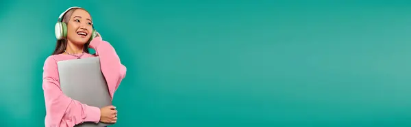 Joyful asian girl in wireless headphones holding laptop while standing on turquoise backdrop, banner — Stock Photo