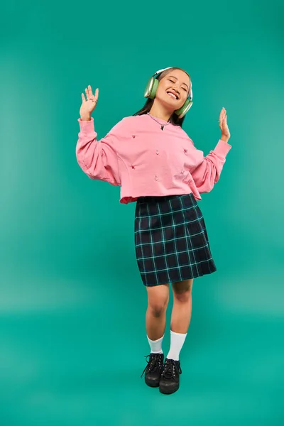 Longitud completa de positivo asiático chica en inalámbrico auriculares bailando en turquesa fondo - foto de stock