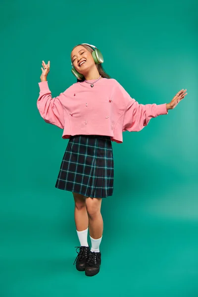 Pleine longueur de positive jeune fille asiatique dans sans fil casque dansant sur fond turquoise — Photo de stock