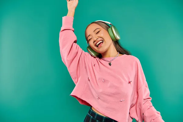 Positive junge asiatische Mädchen in drahtlosen Kopfhörern gestikulieren und tanzen auf türkisfarbenem Hintergrund — Stockfoto