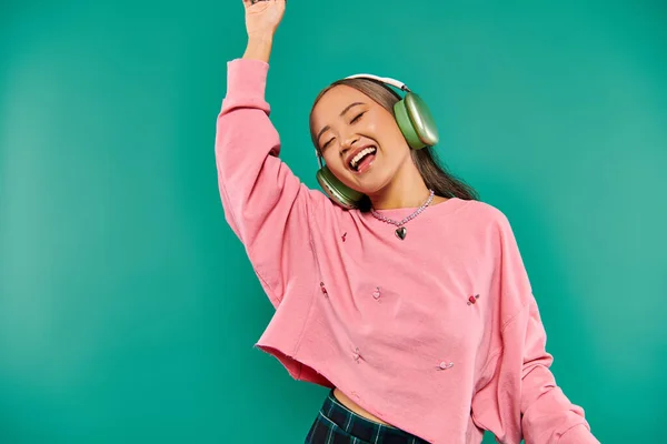 Positivo joven asiático chica en inalámbrico auriculares gesto y bailando en turquesa fondo - foto de stock