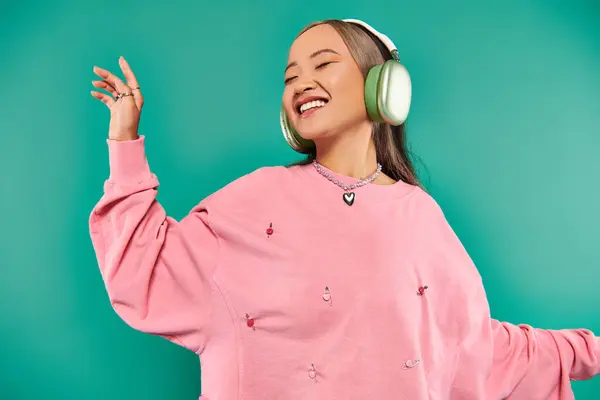 Cheerful young asian girl in wireless headphones gesturing and dancing on turquoise background — Stock Photo